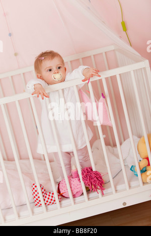 Young girl, 1 year old, with a dummy, standing in her cot and wanting to be picked up Stock Photo