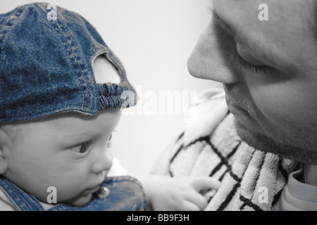 A young baby boy wearing denim dungarees and a blue denim baseball cap backwards sitting with his daddy Stock Photo