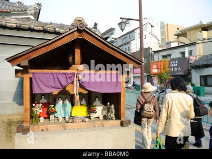 People pass a small shrine-like roadside structure in Ibaraki, a suburb of Osaka, Japan. Such street scenes are very typical. Stock Photo