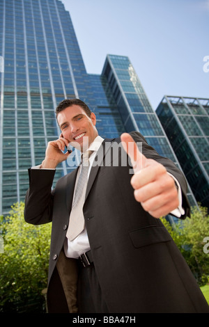 A young male executive in a city surrounding talks on his cell phone and gives a thumbs up Stock Photo
