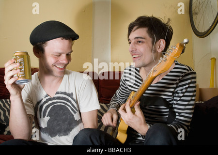 A young couple sitting on a sofa drinking beer and playing guitar Stock Photo