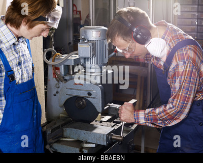 A man using an electric saw in a workshop while a young man looks on Stock Photo