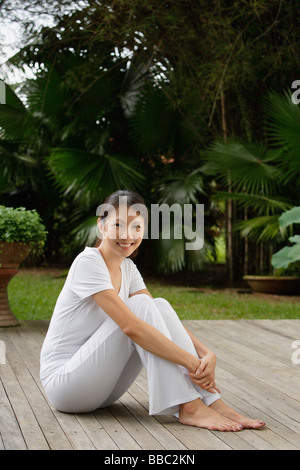 Woman sitting on porch in tropical setting, arms wrapped around legs, looking at camera, smiling Stock Photo