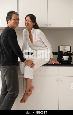 Woman sitting on counter in kitchen with man leaning into her, looking at camera, smiling Stock Photo