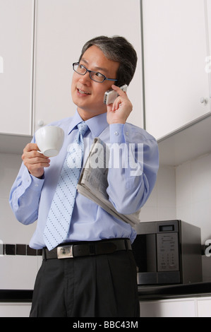 Business Man in kitchen, talking on phone, holding coffee cup and newspaper, multi tasking. Stock Photo