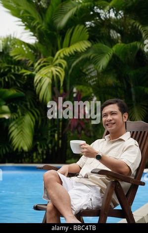 Man sitting by swimming pool, holding a cup of coffee, smiling Stock Photo