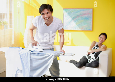 Couple at home, man ironing, woman sitting on sofa behind him Stock Photo