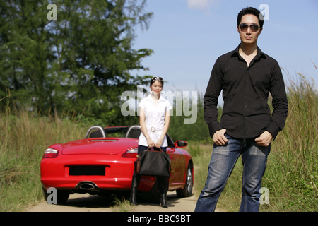 Man wearing sunglasses, facing camera, woman and red sports car in the background Stock Photo
