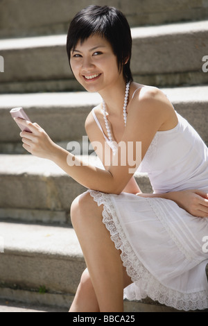 Woman sitting on steps, holding mobile phone, smiling at camera Stock Photo