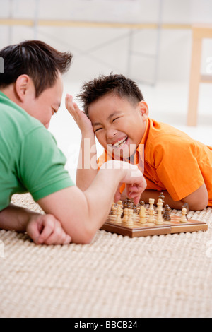 Father and son lying on floor, playing chess, son laughing Stock Photo