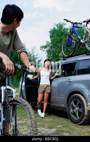 Man on a bike turning to look at woman standing next to car Stock Photo