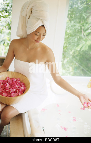 Woman in towel, sitting at edge of bath tub, throwing flower petals into water Stock Photo