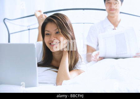 Couple on bed, woman using laptop, man behind her, reading newspaper Stock Photo