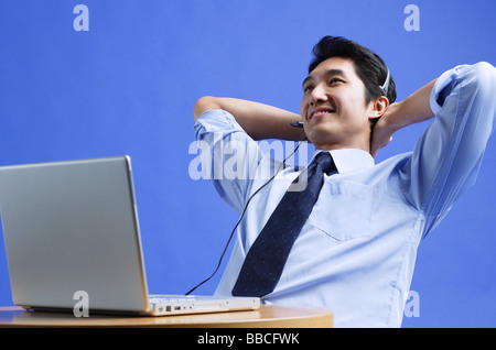 Man wearing headset, using laptop, leaning back, hands behind head Stock Photo
