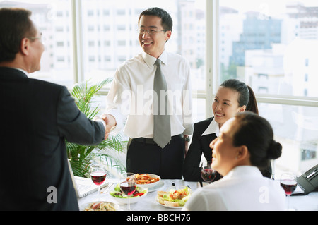 Businessmen shaking hands over lunch table, businesswomen sitting next to them Stock Photo