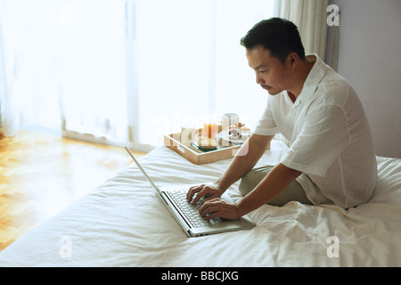 Man sitting on bed, using laptop, breakfast tray next to him Stock Photo