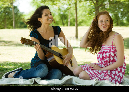 Two young women sitting on blanket outdoors, one playing acoustic guitar Stock Photo