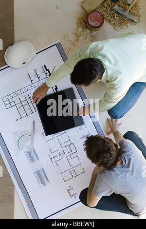 Two men crouching looking over blueprints, using laptop set on floor Stock Photo