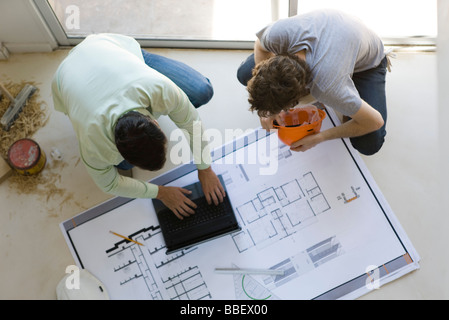 Two men crouching using laptop, looking at blueprint spread out on floor Stock Photo