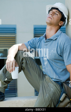 Construction worker sitting on ledge, leaning head back, eyes closed, taking break Stock Photo