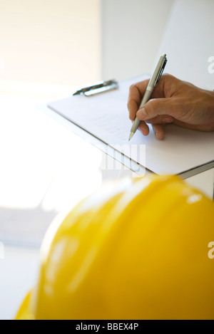 Hand writing notes on clipboard pad, hard hat in foreground, cropped view Stock Photo