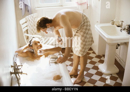 Couple together in bathroom, woman in bathtub, man leaning over her with shaving cream on face Stock Photo