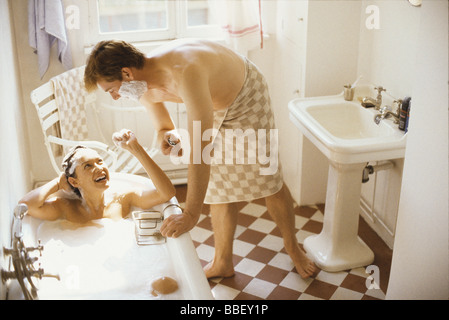 Couple together in bathroom, woman in bathtub, man leaning over her with shaving cream on face Stock Photo