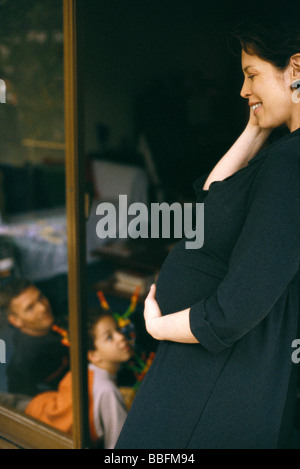 Pregnant woman leaning in doorway, smiling, family in background Stock Photo