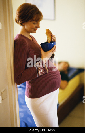 Pregnant woman leaning in doorway, eating croissant Stock Photo