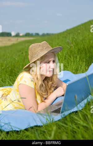 Blond girl wearing a hat, smiling and working on a laptop while lying on a blanket on a meadow Stock Photo