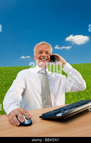 Business concept shot showing an older male executive sitting at a desk using his computer and cell phone in a green field Stock Photo