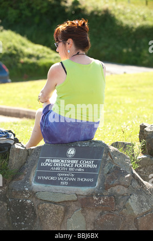 A woman sitting on the signpost marking the start of the Pembrokeshire Coast long distance footpath at Poppit Sands Wales UK Stock Photo