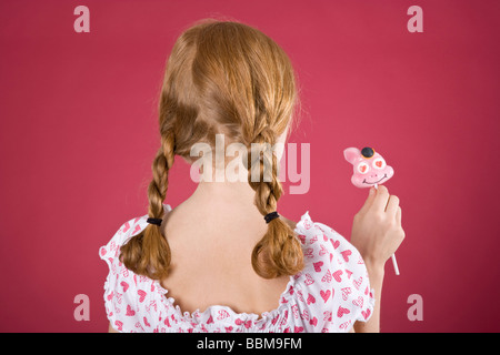 Red-haired girl with braids wearing a summer dress in front of a red backdrop with a lollypop Stock Photo