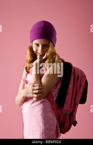 Red-haired girl wearing a violet bonnet and a schoolbag in front of a pink backdrop Stock Photo