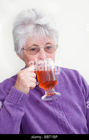 Lonely senior woman looking sad drinking red fruit tea in a glass mug alone at home. England UK Britain Stock Photo