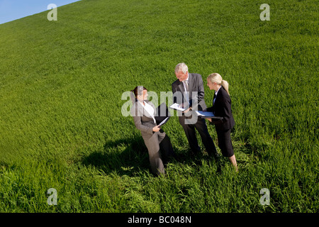 High angle concept shot of a team of three people one man and two women in a green field with a bright blue sky looking at plans Stock Photo