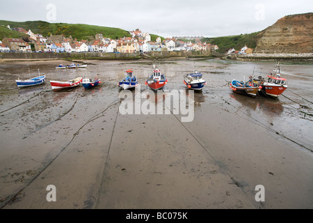 Small tied-up boats in a row left dry at low tide standing on sand with reflections Staithes North Yorkshire England Stock Photo