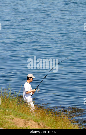 One teenage boy fishing at waters edge with fishing pole hat and white shirt on Cape Cod Canal in Bourne, Massachusetts Stock Photo