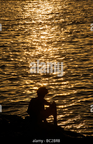 Silhouette of young Cuban man sitting on the rocks by the Malecon. Spikey hbacklit hair Stock Photo