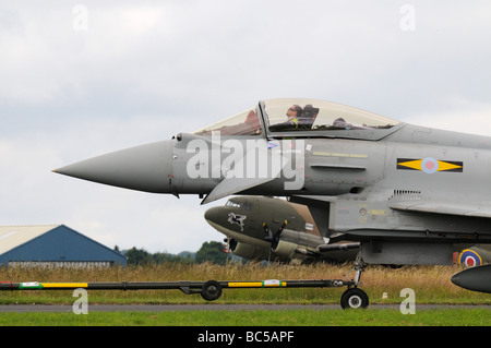 RAF Typhoon Eurofighter is towed along runway past a parked C 47 Dakota whilst crewman rests in cockpit Stock Photo