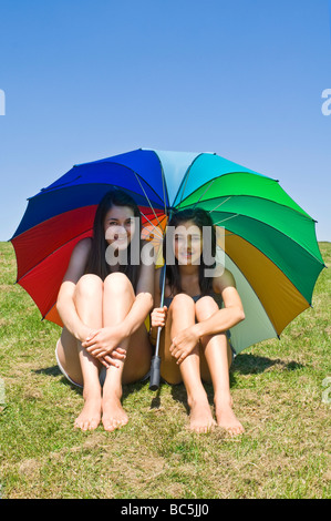 Vertical portrait of two young sisters shielding themselves from the sun underneath a huge colourful umbrella on a summer's day Stock Photo
