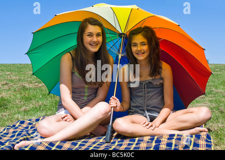 Horizontal portrait of two young sisters shielding themselves from  sun underneath a huge colourful umbrella on a summer's day Stock Photo