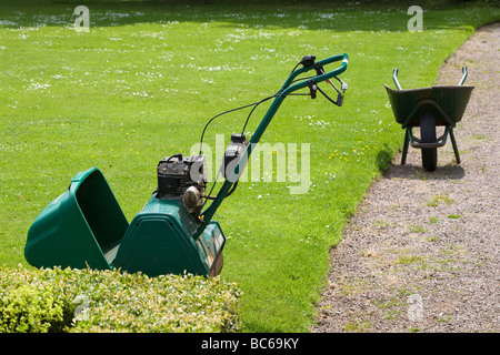 Petrol lawn mower and wheelbarrow sitting on lawn and gravel path Stock Photo