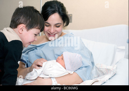 Hispanic mother holds her newborn in the hospital as her three year old looks on. Stock Photo