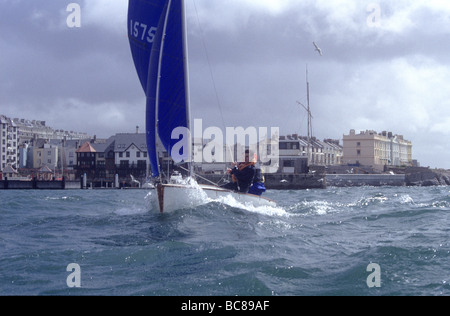 sailing on sea in small white bosan dinghy yacht boat houses building in background Plymouth Devon westcountry Stock Photo