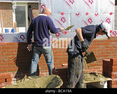 builder / construction worker working on a brick wall cementing bricks in with his apprentice mixing up cement Stock Photo
