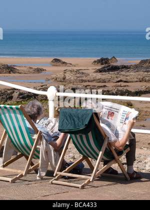 Old couple sat on deck chairs reading newspapers at the beach Stock Photo