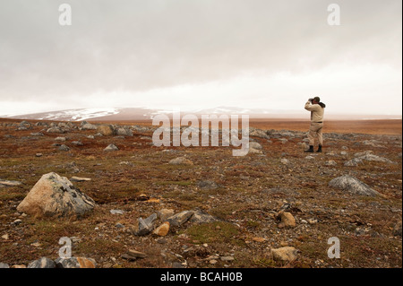 A MAN STANDS IN A FIELD OF ROCKS AND BOULDERS LOOKING THROUGH BINOCULARS Stock Photo