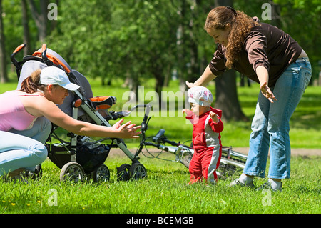 Two women help toddler to do first steps in the park. Green grass and foliage around they. It`s a summer. Baby carriage and bike Stock Photo