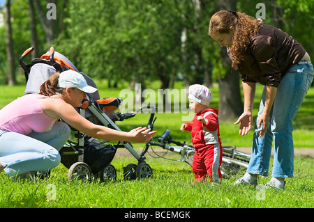 Two women help toddler to do first steps in the park. Green grass and foliage around they. It`s a summer. Baby carriage and bike Stock Photo